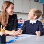 Young female primary school teacher and schoolboy sitting at a table working one on one, looking at each other smiling, front view, close up