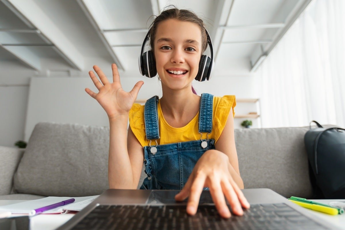 A young girl waving at a laptop screen, she has a smile on her face and is wearing headphones.