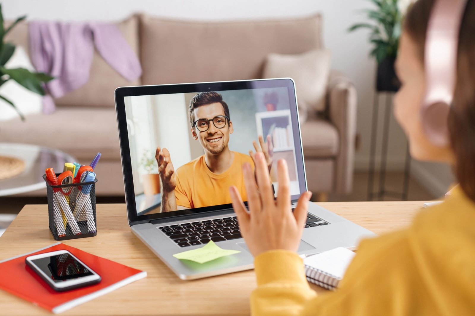 A young girl with pink headphones on having an online lesson through her laptop with a couch in the background.