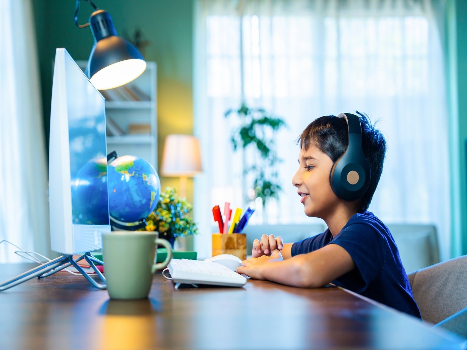 A young indian boy learning online at home using his home computer, with a well lit window in the background and a lap which is on shining down at the computer.