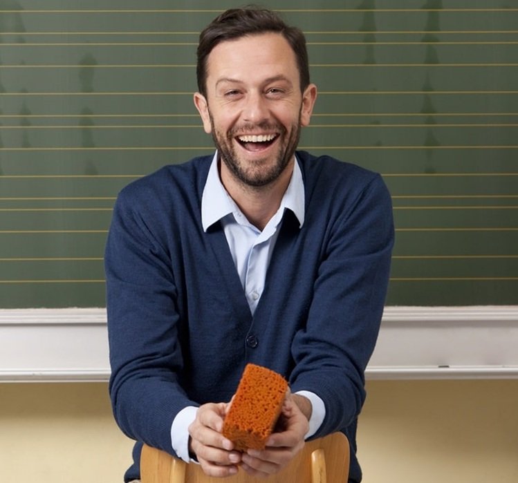 laughing-teacher-in-classroom-at-blackboard-2024-09-17-05-22-54-utc-min A teacher smiling with a red sponge in his hand, who is an online tutor for ATTC Education.