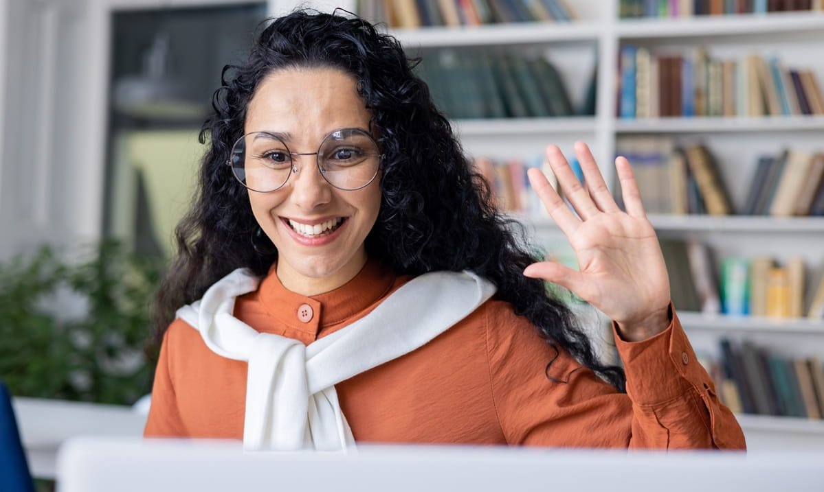 Happy female teacher conducts online class in office, waves at camera and smiles