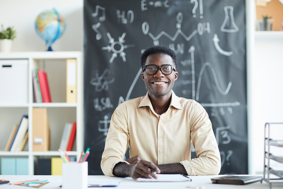 Happy school teacher with toothy smile sitting by desk in classroom with blackboard on background